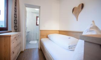 A cozy room with wooden details and a single bed. In the background, a modern bathroom is visible.