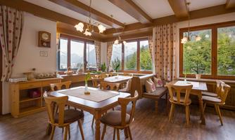 A cozy dining area with wooden furniture and large windows overlooking a green landscape. Bright curtains and natural lighting create an inviting atmosphere.