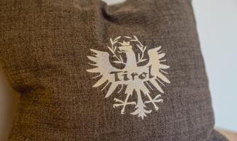 A brown pillow with a white coat of arms. The coat of arms features the inscription "Tirol" and a stylized eagle.
