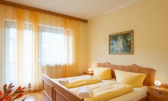 A bright bedroom with a large bed and yellow bedding. The windows are adorned with transparent curtains, and a painting hangs in the room.