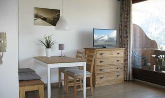 A modern living room with a dining table and chairs. In the background, there is a television and a dresser, as well as large windows with a view outside.