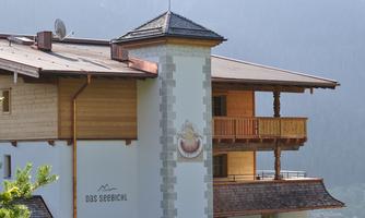 A modern-designed mountain hotel with wooden cladding and a small balcony. In the background, the mountains and a clear sky are visible.