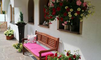 A cozy porch with a wooden bench and colorful flower pots. Hanging planters adorn the wall, and the floor is covered with stone slabs.