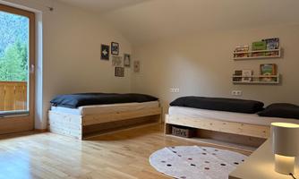 A cozy bedroom with two single beds and a light wood floor. The room has a window overlooking nature and a modern table lamp.