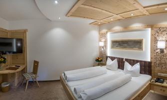 A cozy hotel room with a double bed decorated with white pillows. The walls are made of wood, and there is a flat-screen TV as well as a small table.