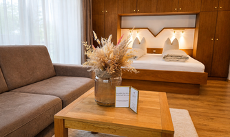 A modern bedroom with a double bed and a cozy couch. On the table, there is a vase with dried flowers and a small card.