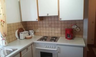 A small, bright kitchen with white cabinets and a tiled wall. On the countertop, there is a coffee machine and there are cooking plates as well as an oven.