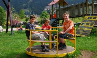 Three children are sitting on a popular piece of outdoor playground equipment. In the background, trees and playground structures can be seen.