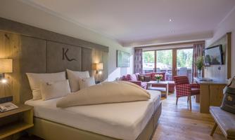 A modern hotel room with a double bed and a cozy seating area. Large windows provide plenty of natural light and a view outside.