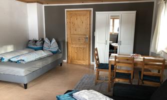 A cozy room with a sofa bed and a wooden dining area. The space has a bright atmosphere with a wooden door and modern decor.