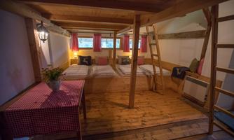 A cozy sleeping room with two beds, a wooden ladder, and beautiful window shutters. A table with a red-and-white tablecloth and a flower vase creates an inviting atmosphere.