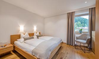 A modern bedroom with a double bed and light wooden floor. Daylight streams through the window onto a chair and plants.