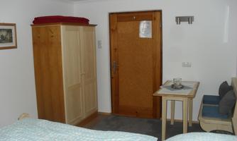 A simply furnished room with a bed, a wardrobe, and a table. The wall is white and a door leads outside.