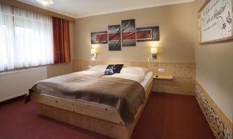 A cozy bedroom with a large bed and wooden details. Bright windows create an inviting atmosphere.