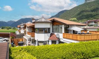A modern house with a large terrace and green hedges. In the background, mountains and a blue sky are visible.