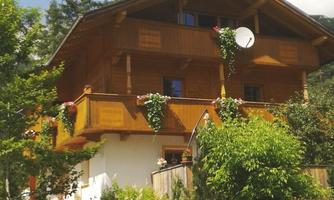 A charming wooden house surrounded by trees and plants. The sky is blue with some clouds.