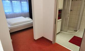 A quiet hallway with a bedroom on the left side and a bathroom on the right side. The floor is red and the bedroom has windows with curtains.