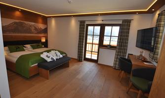 A cozy hotel room with a large bed, warm wood tones, and modern furniture. There is a balcony with a view and a desk for working.