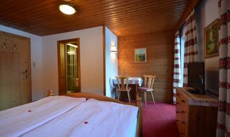 A cozy room with a double bed and a wooden table. The walls are partially made of wood, and there is a shower in the background.