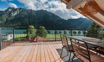 A spacious terrace with a table and chairs, offering a stunning view of mountains and a lake. The sky is partly cloudy and the landscape is green and inviting.