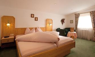 A cozy bedroom with a large double bed and wooden details. The room features an armchair and a small table next to a window with curtains.