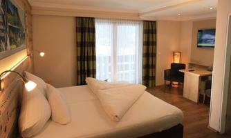 A modern hotel room with a double bed and bright interior. Large windows let in plenty of light, and there is a small desk in the corner.