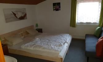 A cozy bedroom with a double bed and a sofa bed. The walls are light, and there is plenty of daylight through the window with curtains.