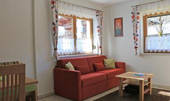 A cozy living room with a red sofa and a small table. The windows let in a lot of daylight and the walls are brightly decorated.