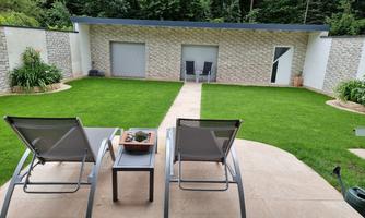 A well-maintained garden with green lawn and a modern terrace. In the background, a garden shed and two chairs can be seen.