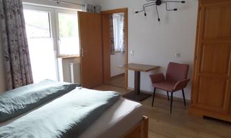 A cozy bedroom with a double bed, a table, and an armchair. Bright walls and large windows create a friendly atmosphere.