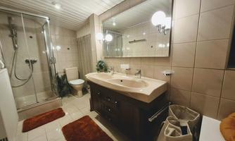 A modern bathroom with a shower, a large mirror, and a sink. The walls are tiled, and there are some decorative elements like plants and bath mats.