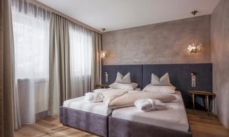 A modern bedroom with two beds and elegant pillows. The room has large windows and a bright, cozy atmosphere.