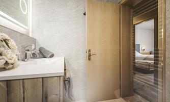 A modern bathroom with a wooden washbasin and a large mirror. An open door leads to a bedroom in the background.