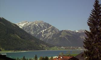A beautiful view of mountains and a clear lake. In the foreground, some roofs and trees can be seen.