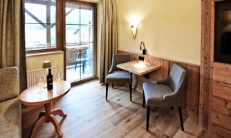 A cozy living space with a small table and two chairs. Large windows provide a view outside, and the decor is in warm wood and earth tones.