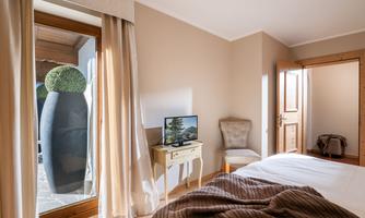 A cozy bedroom with a large window and a balcony. The decor is modern and bright, featuring a comfortable bed and a chair.