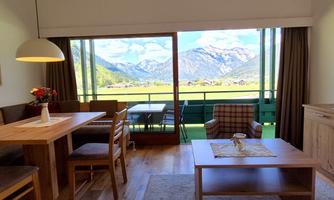 A cozy living room with wooden furniture and a large window. The view shows the impressive mountain landscape.
