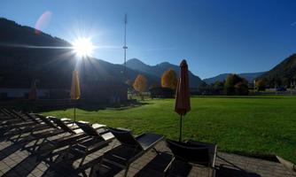 A sunny day in a green square with loungers and sun umbrellas. In the background, there are mountains and a clear sky.