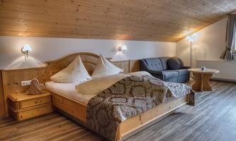 A cozy bedroom with a double bed and a comfortable seating area. The wooden ceiling and warm light create an inviting atmosphere.