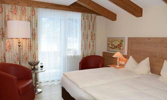 A cozy bedroom with two single beds and wooden ceilings. Red chairs and a window with curtains create an inviting atmosphere.