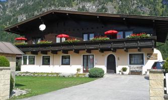 A charming alpine house with a large balcony and red sun umbrellas. The surroundings are green and welcoming, with a view of the mountains.