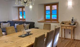 A cozy dining area with a large wooden table and comfortable chairs. An elegant ceiling lamp and bright windows create an inviting atmosphere.