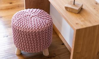 A round, pink stool with a knitted pattern stands next to a wooden cabinet. The stool has three light legs and gives the room a cozy atmosphere.