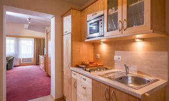 A modern kitchen with wooden cabinets and stainless steel appliances. In the background, a cozy room with a red carpet is visible.