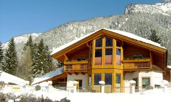 A charming wooden house in winter, surrounded by snow-covered mountains. The sun shines on the large window front of the house.
