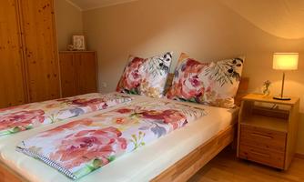 A cozy bedroom with a double bed and blooming pillows. Next to the bed is a bedside lamp and a wooden wardrobe in the background.