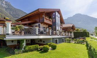 A beautiful wooden house with a balcony and many plants. In the background, majestic mountains and a clear sky can be seen.