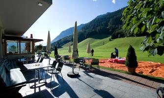 A terrace with tables and chairs, surrounded by green meadows and mountains. In the background, people can be seen with a parachute.