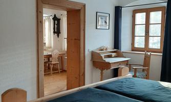 A cozy bedroom with a wooden bed and a large window. In the background, you can see a table and chairs in an adjoining room.