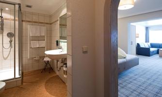 A modern bathroom with shower and sink, next to a living room with comfortable furniture. The room is bright and inviting.